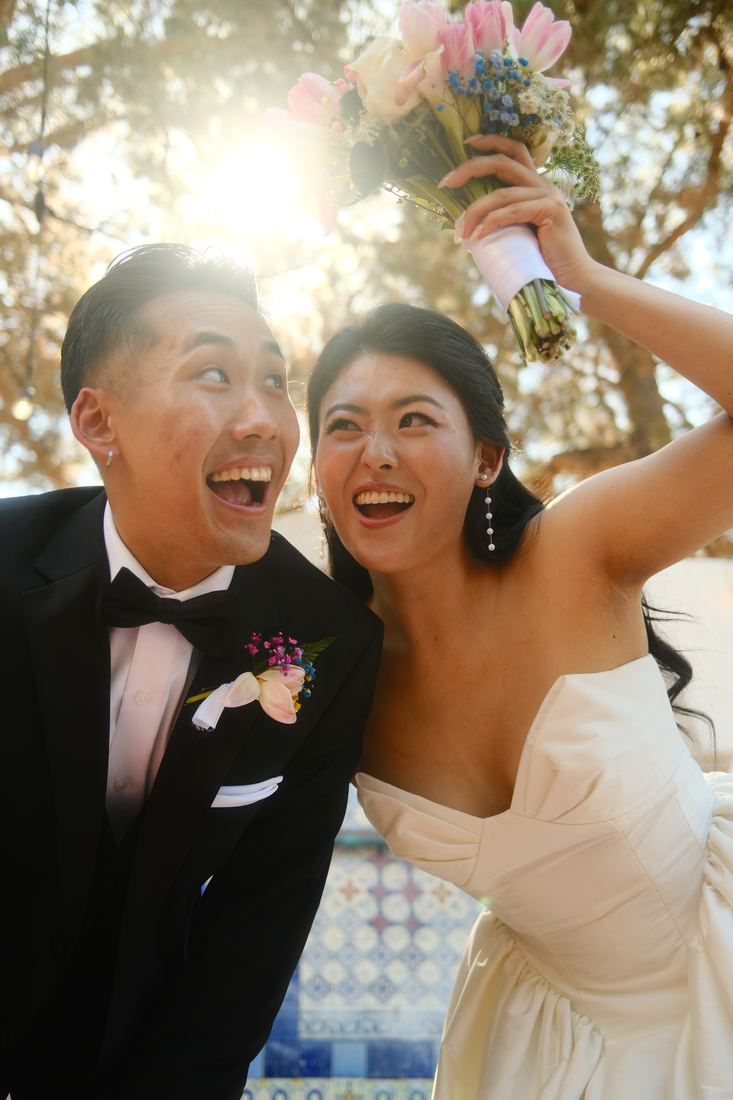 Groom and bride laughing, bouquet raised into the sun